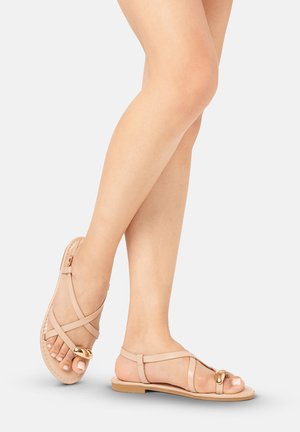 Piernas cruzadas con sandalias planas beige con tiras cruzadas y detalle de hebilla dorada sobre un fondo claro y liso.