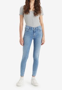 Kvinna i ljusblå skinny jeans, en grå kortärmad skjorta med knappar, vita strumpor och vita sneakers, stående mot en vit bakgrund.