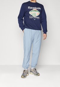 Marineblå sweatshirt med "Reebok Court Classic" grafisk tryck, ljusblå veckade byxor och grå träningsskor med gula detaljer.
