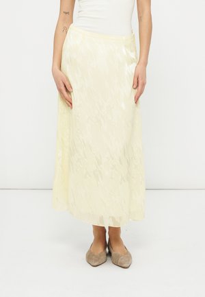 Ženska, oblečena v svetlečo kremno midi krilo z visokim pasom, belo brezrokavico in bež koničaste balerinke, stoji pred enobarvno belo ozadje.