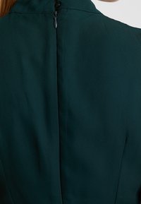Dunkelgrünes Kleid mit hohem Halsausschnitt und einem Reißverschluss im Rücken. Der Stoff wirkt glatt und leicht strukturiert mit einer taillierten Passform.