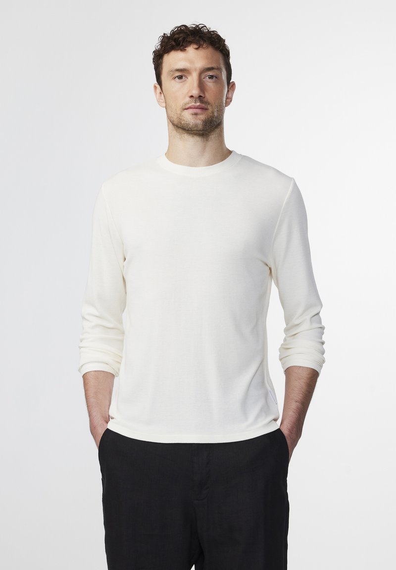 Long-sleeve cream-colored shirt in a lightweight, textured fabric, featuring a crew neck and fitted sleeves, worn with dark trousers.
