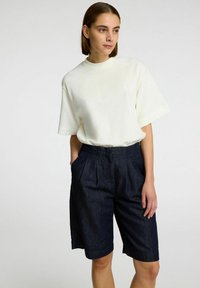 Chemise blanche à manches courtes avec col rond, associée à un short en denim bleu foncé présentant des plis et des poches latérales, offrant une coupe décontractée.