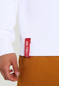 Witte sweatshirt met geribbelde manchetten, voorzien van een rood geweven label aan de zoom. Gecombineerd met mosterdkleurige broek.