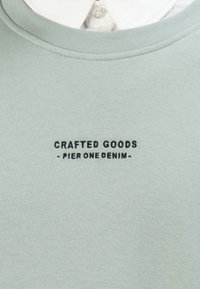 Lichtgrijs T-shirt met ronde hals en zwarte tekst "CRAFTED GOODS - PIER ONE DENIM -" op de borst, gedragen over een witte blouse.