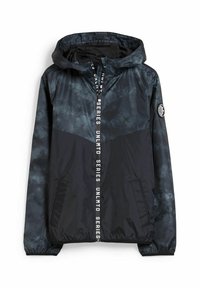 Chaqueta cortavientos de tie-dye en negro y gris con capucha, cierre de cremallera blanca y bolsillos laterales; presenta un parche de logo en la manga.