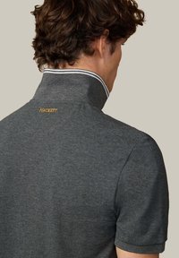 Sivý polo tričko s vysokým golierom, s bielym pruhovaným akcentom a vyšitým logom "HACKETT" v zlatej farbe na chrbte. Mäkká textúra.