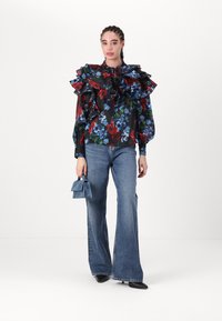Floral blouse with ruffled sleeves in black, red, and blue, paired with flared blue jeans. Model holds a small blue handbag and wears black shoes.