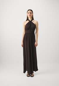 Robe halter longue et fluide en tissu marron foncé avec corsage plissé et taille élastique. Le mannequin porte des sandales noires et se tient sur un fond neutre.