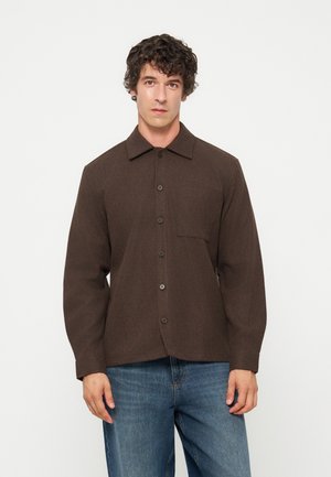 Mann trägt ein braunes Hemd mit Knöpfen und langen Ärmeln und blaue Jeans und steht vor einem schlichten weißen Hintergrund.