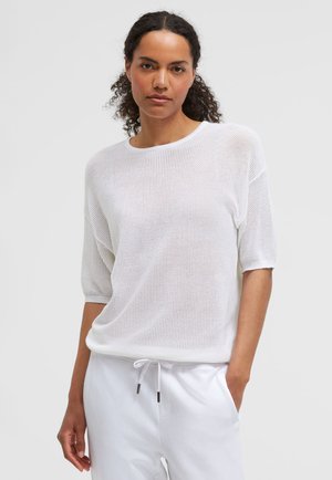 Frau mit lockigem Haar, die ein weißes kurzärmeliges Stricktop und weiße Hosen mit Kordelzug trägt, steht mit einer Hand in der Tasche.