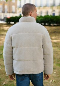 Jachetă puffer bej din fleece, cu guler înalt și panouri orizontale cusute. Purtați-o cu blugi albaștri, evidențiind o textură moale și un croi lejer.