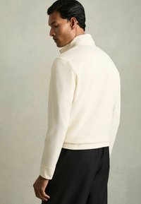 Weißer Pullover mit hohem Kragen und strukturiertem Stoff, mit langen Ärmeln und taillierter Passform, dazu schwarze Hose.