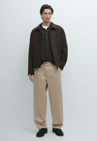 Giacca marrone con bottoni sopra una polo grigio scuro, abbinata a pantaloni beige a gamba larga e scarpe nere. Tessuto liscio con colori neutri.