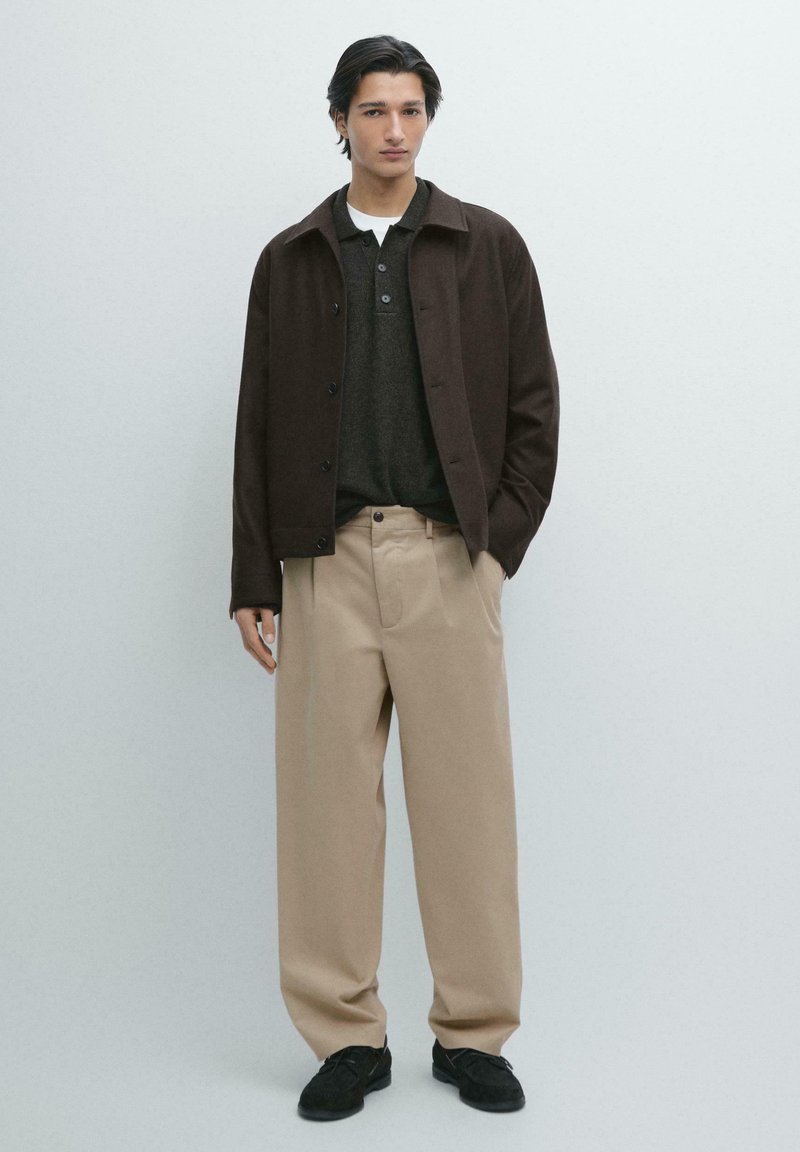 Giacca marrone con bottoni sopra una polo grigio scuro, abbinata a pantaloni beige a gamba larga e scarpe nere. Tessuto liscio con colori neutri.