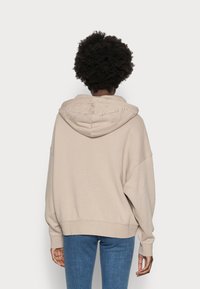 Beżowa bluza z dużym kapturem i haftowanym logo "ESPRIT" na plecach. Ma luźny krój oraz ściągacze przy mankietach i dolnym brzegu. Szerokie, luźne rękawy.