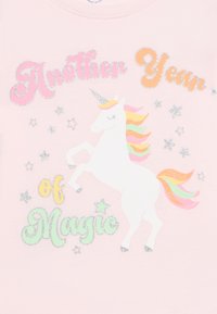 Λευκός μονόκερος με ουράνιο τόξο και λαμπερά ασημένια αστέρια, περιτριγυρισμένος από πολύχρωμο κείμενο "Άλλος ένας χρόνος μαγείας" σε ανοιχτό ροζ ύφασμα.