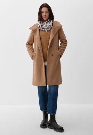 Frau trägt einen Kamelmantel, braunen Pullover, gemusterten Schal, blaue Jeans und schwarze Stiefel, steht mit den Händen in den Manteltaschen.