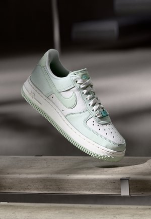 Buty Nike Air Force 1 ze skóry w jasnym zielonym i białym kolorze, unoszące się nad betonową powierzchnią z widocznymi szwami i sznurowadłami.