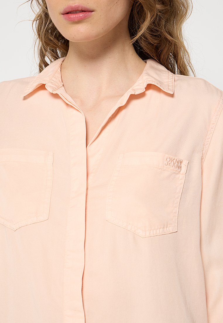 Camisa de botones de color durazno claro, hecha de una tela suave; cuenta con un cuello, dos bolsillos en el pecho y un bordado sutil del logo en el bolsillo izquierdo.