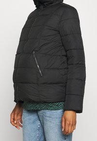 Chaqueta acolchada negra con cierre frontal, bolsillos laterales y capucha. Textura acolchada con un ajuste estándar, llevada sobre una parte superior con patrón.
