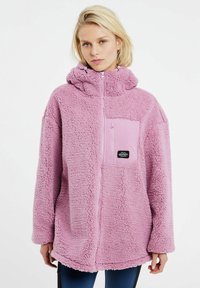 Giacca in pile rosa con cappuccio, dotata di zip frontale a metà, tasca frontale in tessuto rosa e patch logo nero sulla tasca.