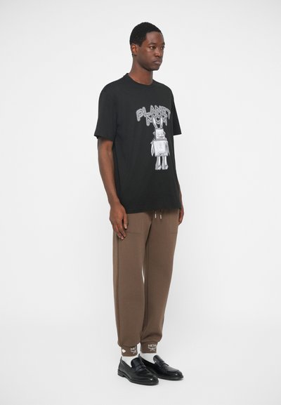 Crna majica s bijelim grafičkim printom koja sadrži natpis "PLANET MCM" i robota. Uske smeđe trenirke s bočnim džepovima i bijelim logotipom.