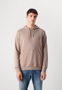 Jonge man in een beige hoodie met een voorzak en een blauwe spijkerbroek, staand tegen een effen lichte achtergrond.