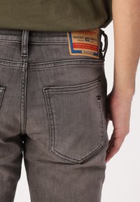 Gray denim jeans with a close-fitting design, featuring two back pockets and a woven label with a blue and orange pattern.