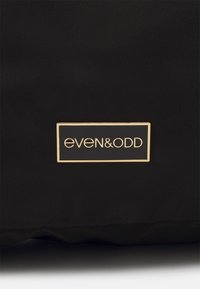Černá strukturovaná taška s obdélníkovým štítkem se zlatými akcenty, na kterém je bílé písmo s názvem značky "even&odd".