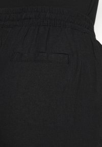 Pantalon noir avec une taille élastique, doté d'une petite poche plaquée à l'arrière. Confectionné dans un tissu doux et lisse avec une finition mate.