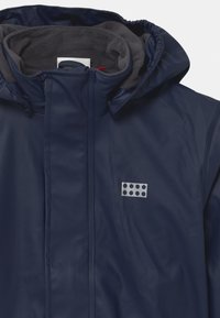 Navy waterproof jacket with a soft gray fleece lining, high collar, zip closure, and a silver logo patch featuring six circular cutouts.