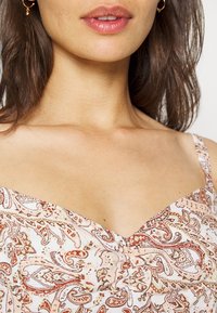 Žena se středně hnědými vlasy, která nosí zlaté kruhové náušnice a béžový top bez rukávů s paisley vzorem a řaseným předním dílem.