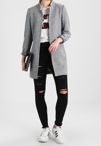 Grå, lång kappa med rak skärning, bärs över en vit t-shirt med grafiskt tryck. Svarta slitna skinny jeans och vita sneakers med svarta ränder.