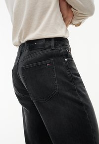 Άτομο που φοράει μαύρα τζιν Tommy Hilfiger και μπεζ μακρυμάνικο πουκάμισο, το χέρι ακουμπάει στη μέση, λευκό φόντο.