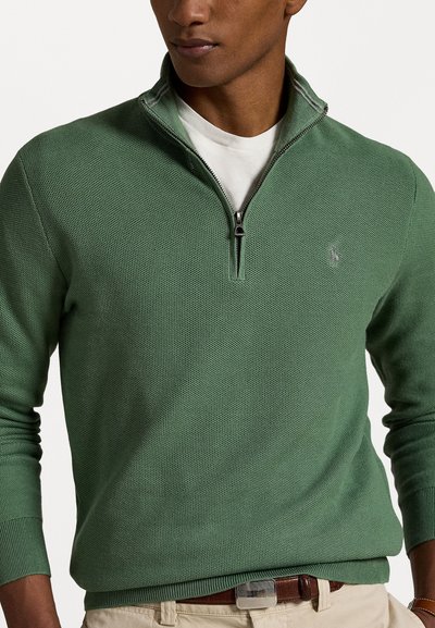 Green knit pullover with a zippered collar. Features ribbed cuffs and hem, and a small logo on the chest. Layered over a white shirt.