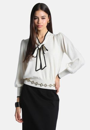 Femme portant un chemisier blanc à manches longues avec un nœud bordé de noir et un détail de chaîne dorée, associé à une jupe noire et des boucles d'oreilles dorées.