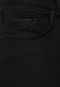 Kieszonka czarnych dżinsów denim z subtelnym wytłoczonym logo w kształcie tarczy i widocznymi detalami przeszycia na teksturowanym materiale.