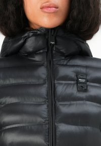 Čierna puffa bunda s vysokým golierom, disponujúca zipsovým zapínaním a horizontálnym prešívaním. Odkaz na logo je viditeľný na hornej ľavej strane.