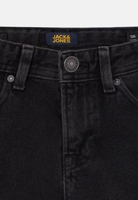 Dettaglio della vita dei jeans in denim nero con chiusura a bottone, passanti per cintura e un'etichetta gialla del marchio Jack & Jones all'interno della vita.