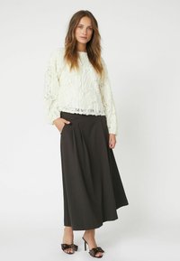 Weißer strukturierter Pullover mit Rüschendesign, kombiniert mit einem langen schwarzen Faltenrock und schwarzen Sandalen mit Absatz, die mit Schleifenakzenten verziert sind.
