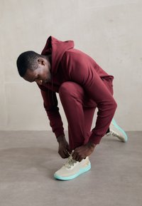 Sportowe sneakersy w odcieniu beżowym z teksturowaną siateczkową cholewką i mięta zieloną podeszwą. Osoba w burgundowym dresie wiąże sznurowadła.