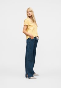 Γυναίκα με κίτρινο κοντομάνικο πουκάμισο και φαρδιά μπλε τζιν με πλαϊνές σκισμές, με τα χέρια στις τσέπες, κοιτάζοντας προς τα αριστερά, φορώντας λευκά μυτερά τακούνια.