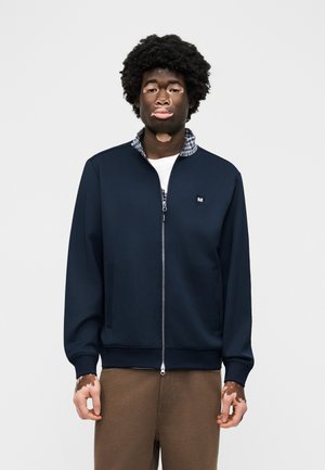 Mladý muž s vitiligom v námorníckej modrej bunde na zips, oblečený cez kockovanú košeľu s golierom a hnedé nohavice, stojaci na bielom pozadí.