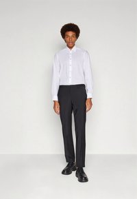 Weißes langärmliges Hemd mit Knopfverschluss, kombiniert mit schwarzen, maßgeschneiderten Hosen und schwarzen eleganten Schuhen, stehend vor einem neutralen Hintergrund.