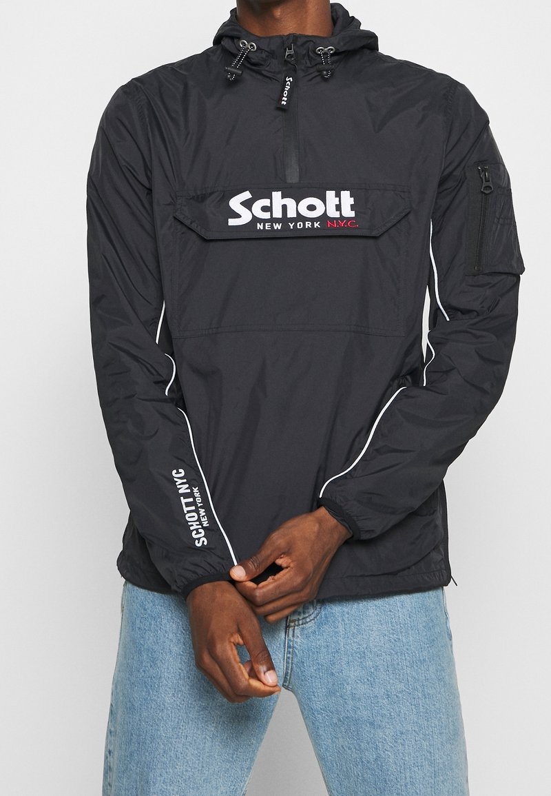 Coupe-vent noir Schott New York avec capuche, fermeture éclair à l'avant, poche sur la manche et texte du logo blanc, porté avec un jean bleu clair.