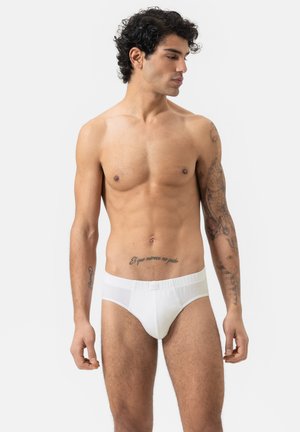 Weiße Baumwollslips mit elastischem Bund, tailliertem Design und glatter Textur. Das Modell zeigt ein Profil von der Seite vor einem schlichten Hintergrund.