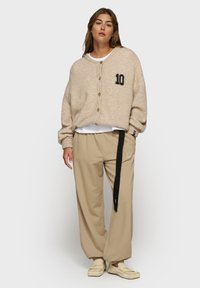 Mujer vistiendo un cárdigan beige abotonado con "10" en el pecho, pantalones beige sueltos con una correa negra, camiseta blanca debajo y mocasines crema.