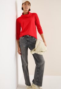 Rotes, langärmeliges Rollkragenoberteil, graue Jeans, hellbeige Kappe und Sneakers. Das Outfit hat eine entspannte Passform mit minimalen Details.