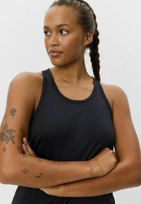 Čierna športová tielko s výstrihom do lodičky a dizajnom s otvorenými chrbtom. Mäkká tkanina s hladkým povrchom. Na rukách sú rôzne tetovania.